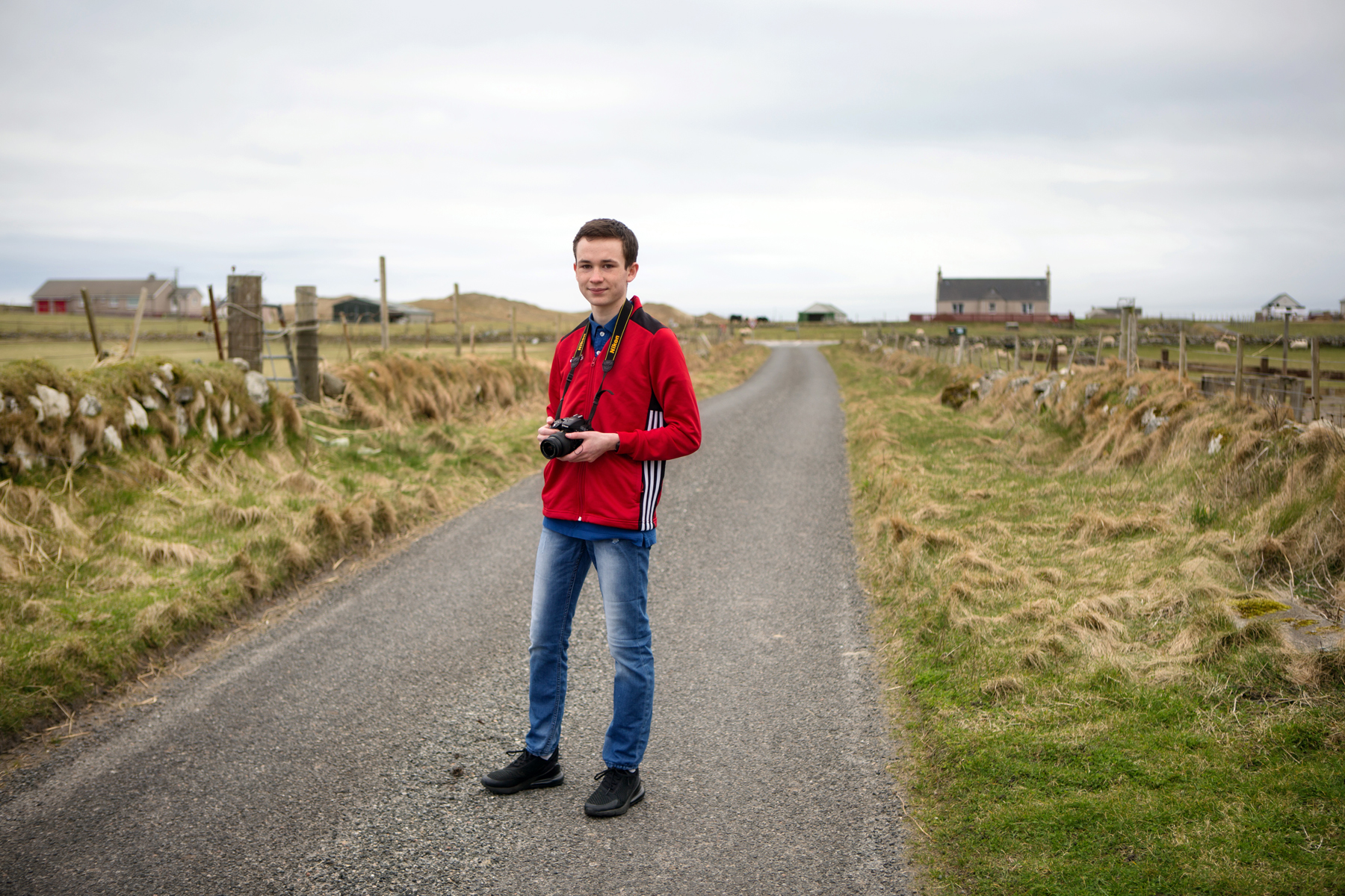 A teenage boy in a red jacket and blue jeans stands on a single track road, with grass either side